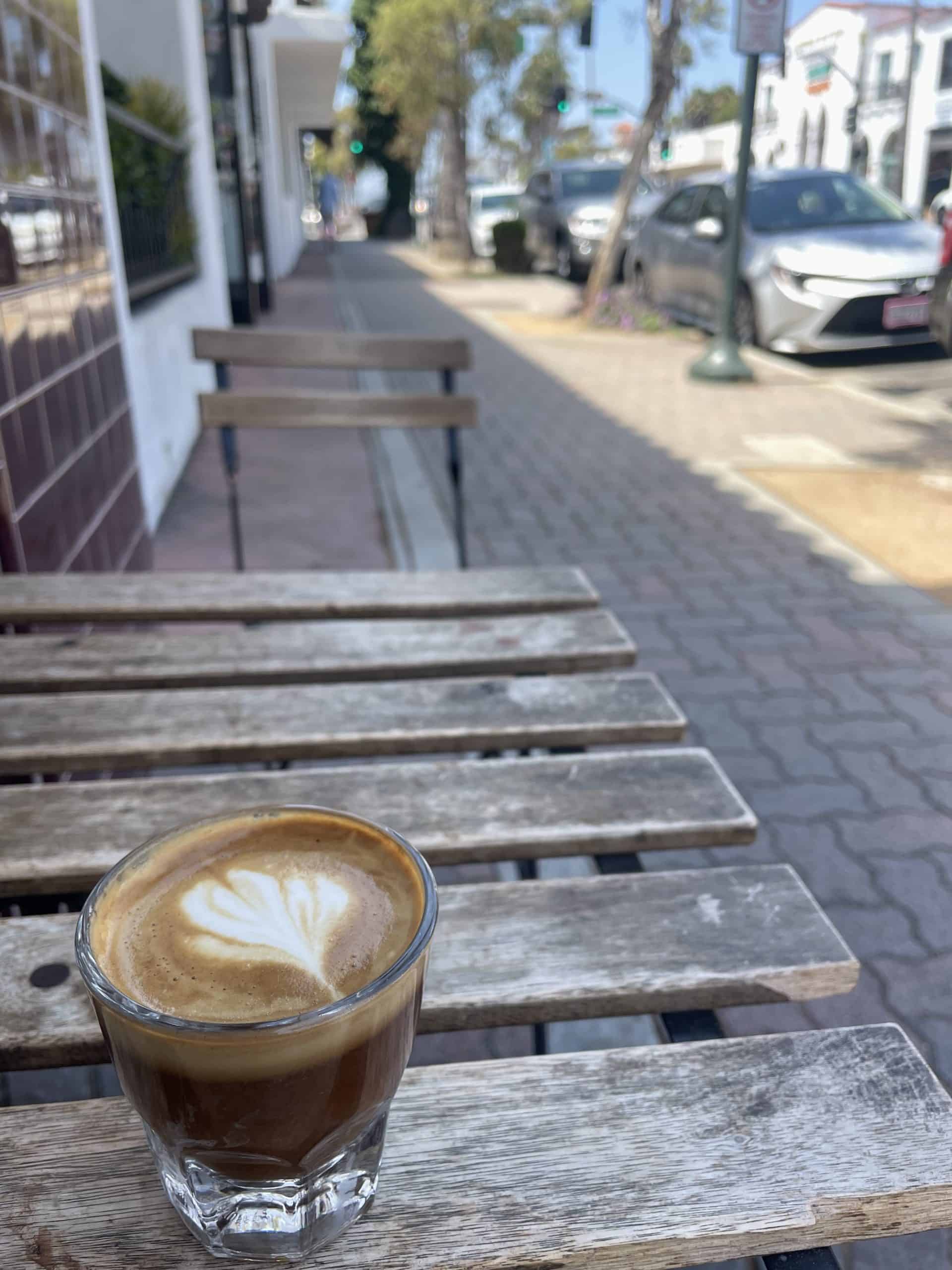 Cortado in a glass cup on a wooden table on a brick pavement in downtown San Clemente, CA