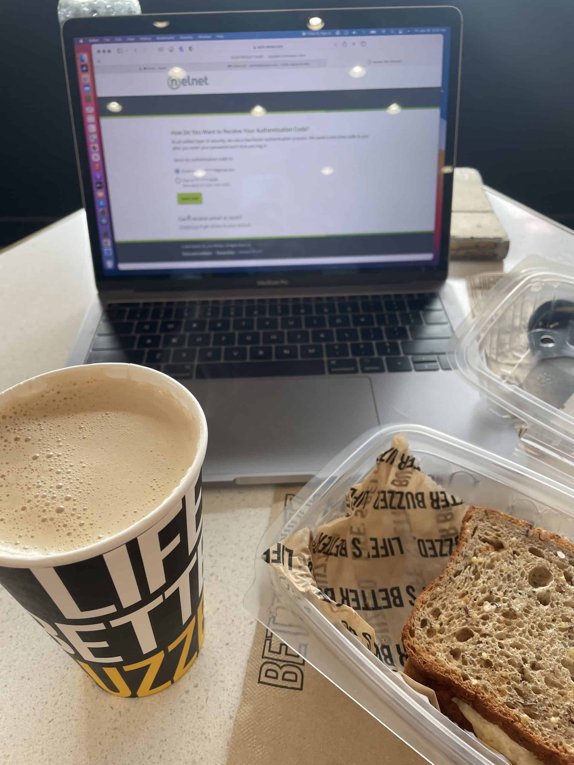 coffee in a paper cup with a sandwich on the right in a plastic container sitting on a table with a laptop open