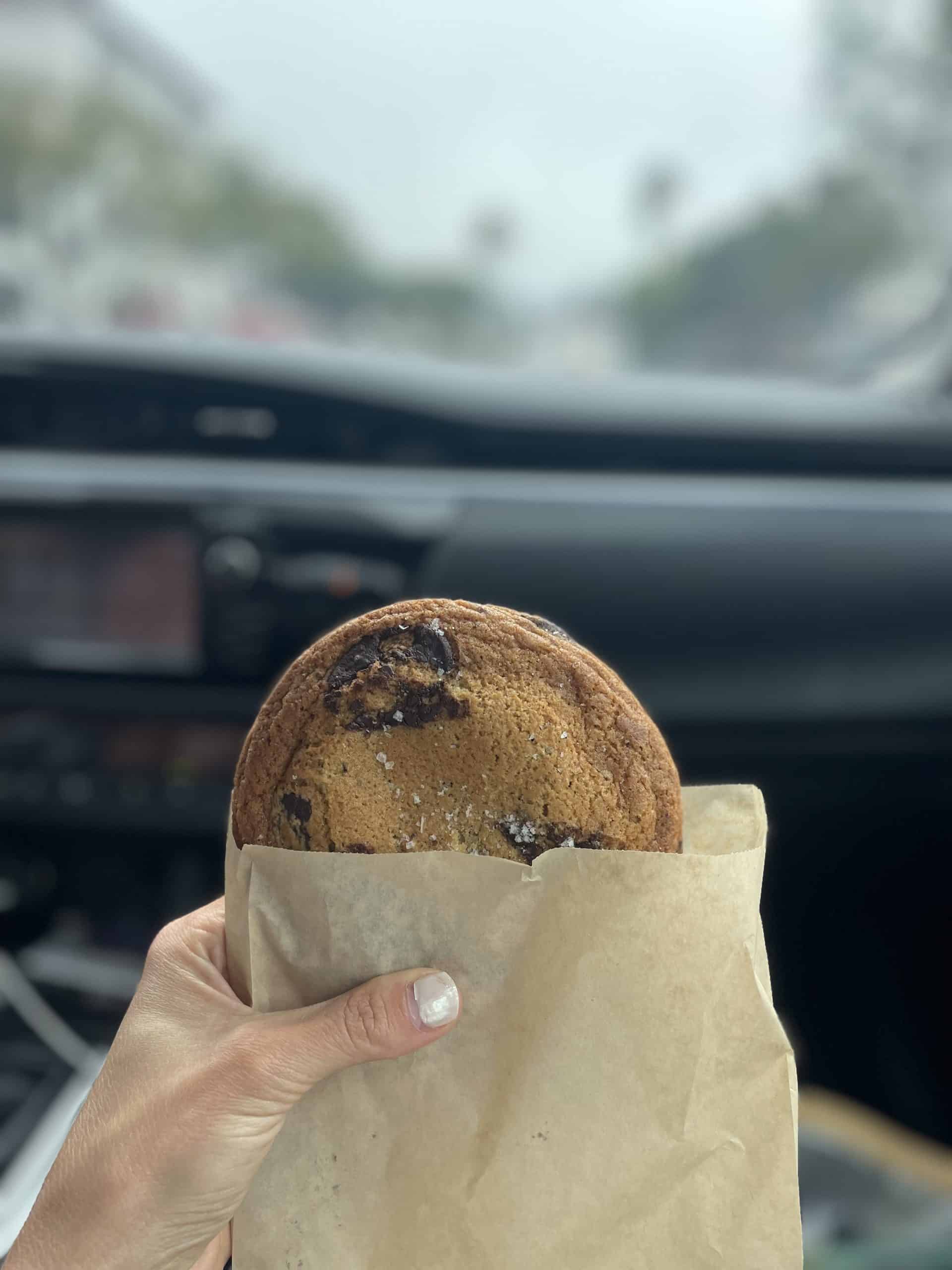 chocolate chip cookie in a paper bag being held with my hand in a car, my nails are painted a glossy white, the background is blurry