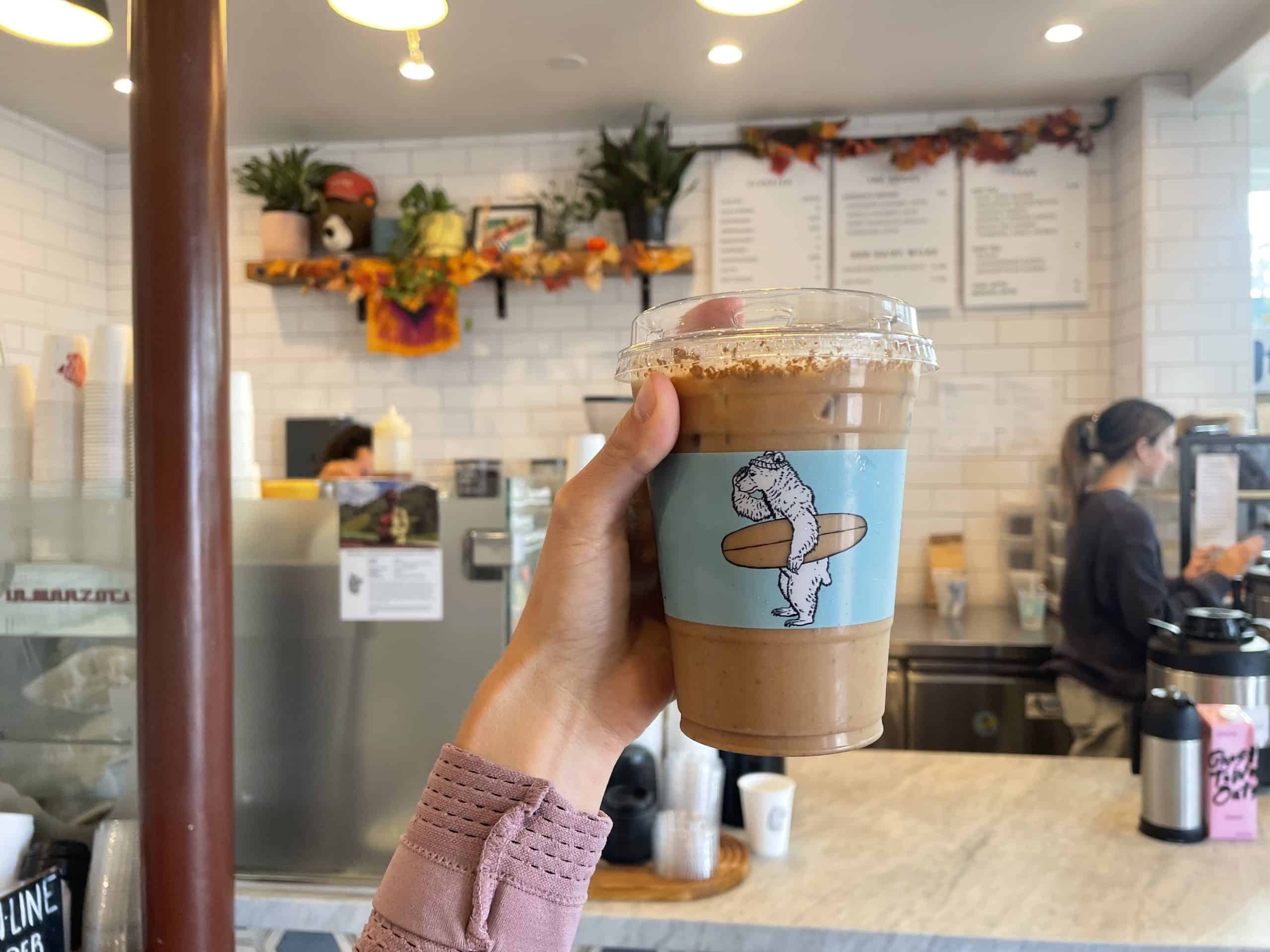 Iced chai latte being held with a hand with a logo on the cup that has a blue stripe and a bear holding a surfboard