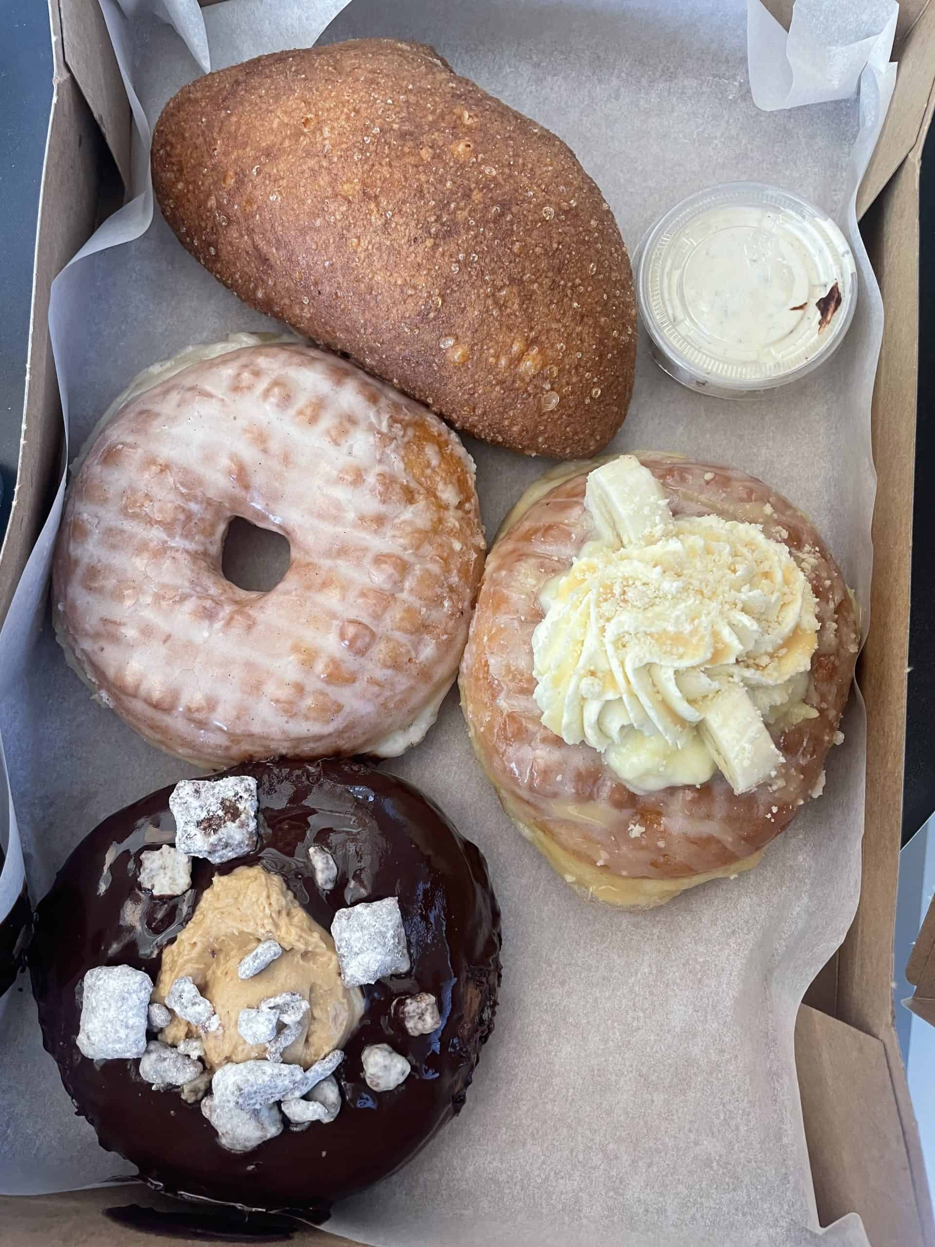 four donuts in a box, one of them with glaze, one with whip cream on top and one with a scoop of peanut butter and puppy chow on top