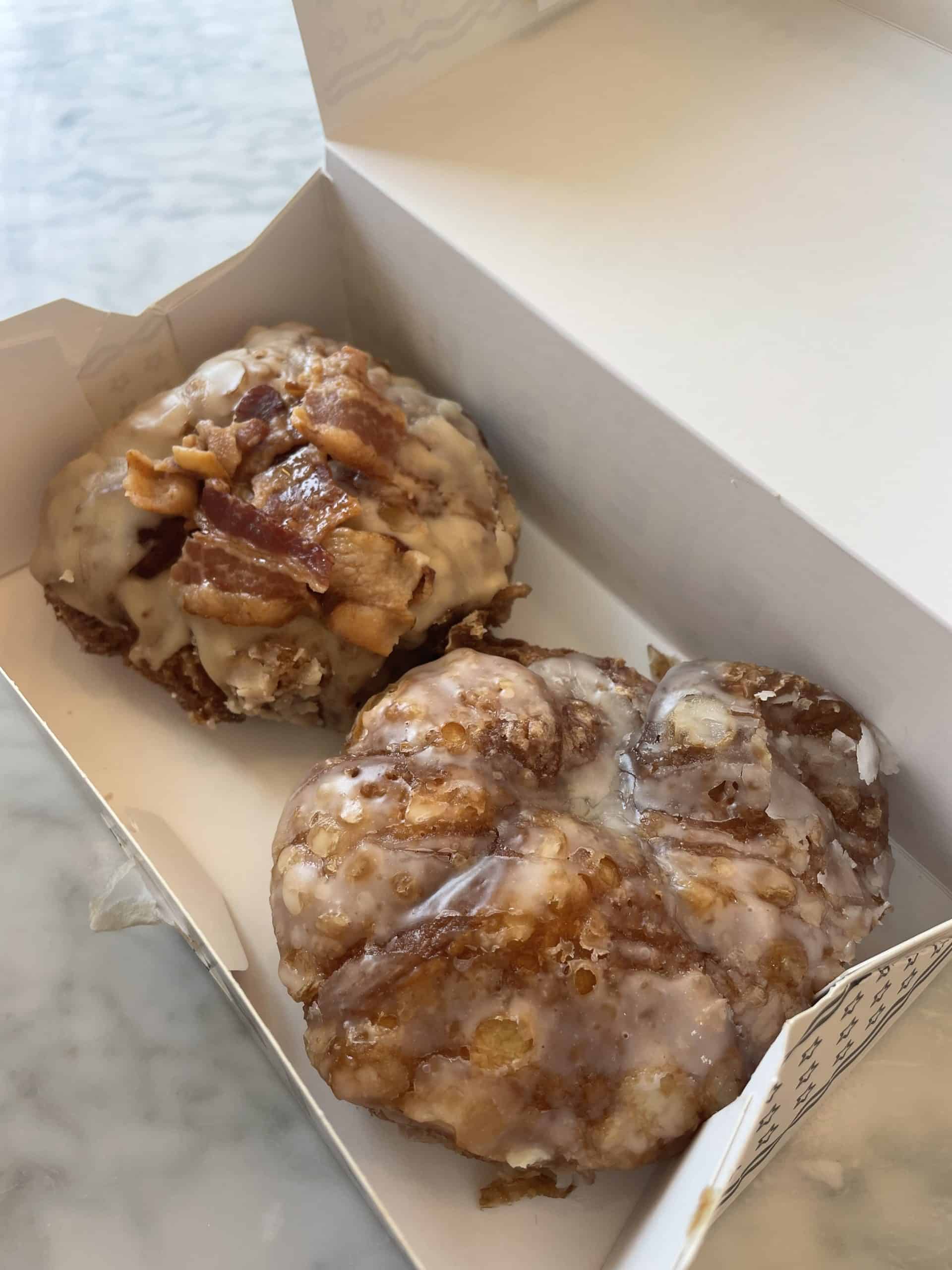 a white box with two thick layered donuts, one glaze and one with bacon on top
