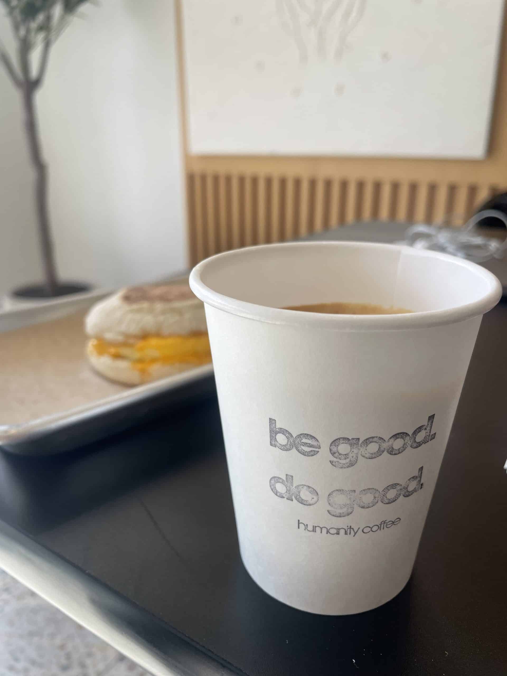 a paper cup that says 'be good do good humanity coffee' with an egg English muffin in the background