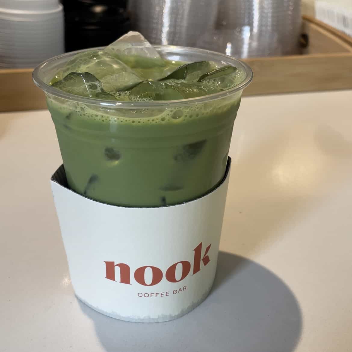 an iced matcha latte in a plastic cup with a cardboard ring around it that says 'nook coffee bar'