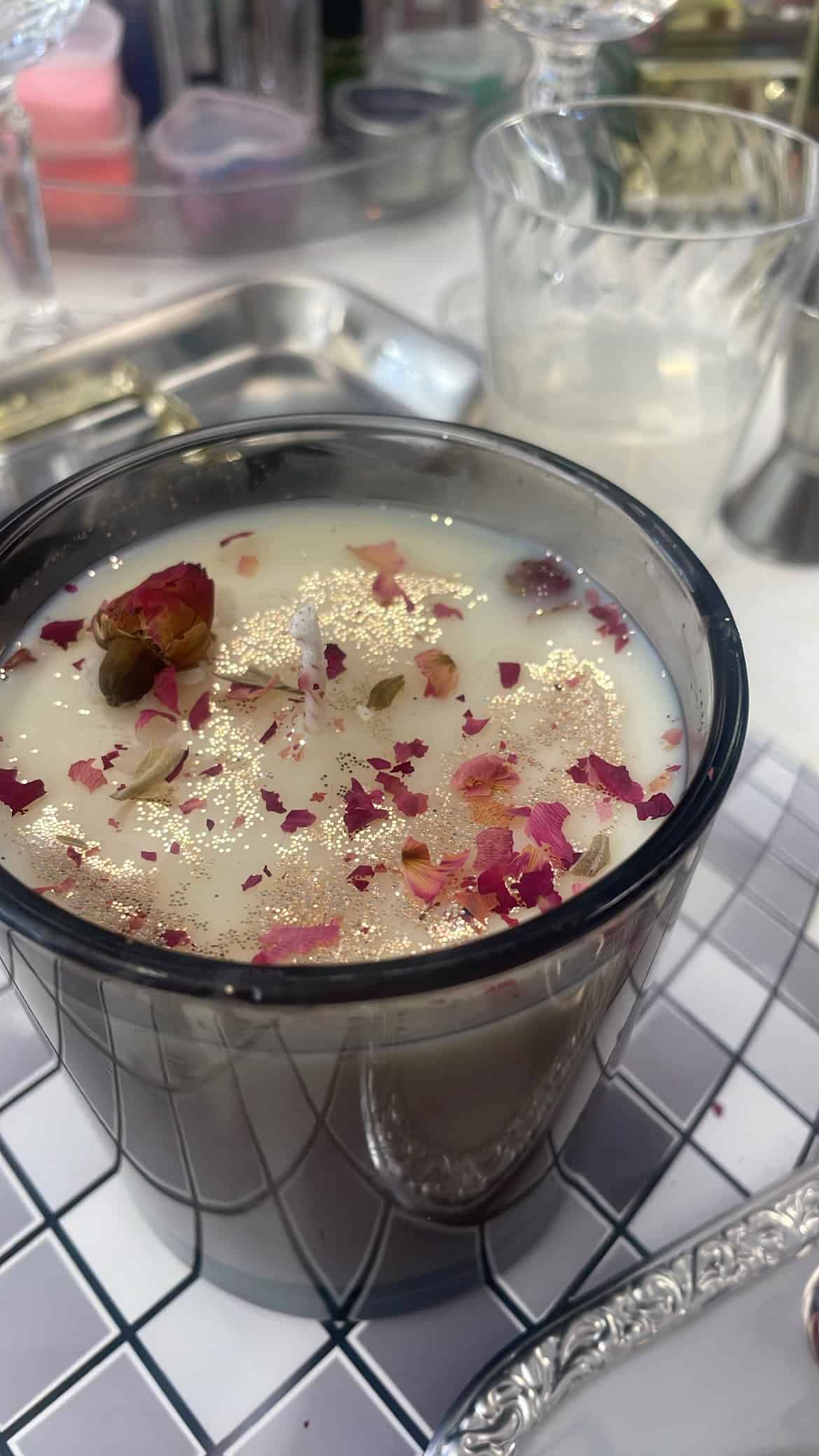 a candle in a dark clear vessel with gold glitter and dried pink rose petals on top