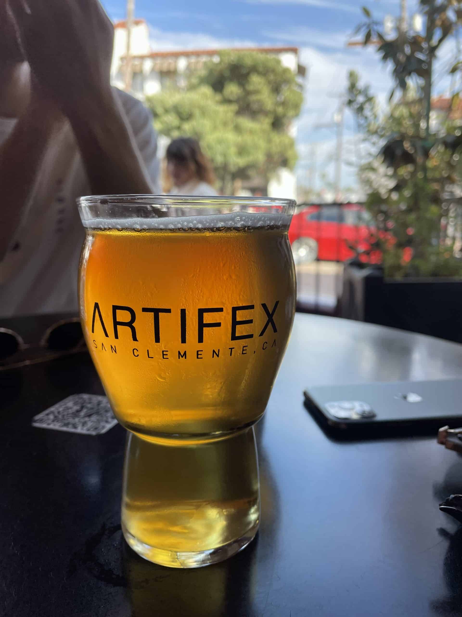 a beer in a clear cup that says 'Artifex San Clemente, CA' on a black table with a blurred background