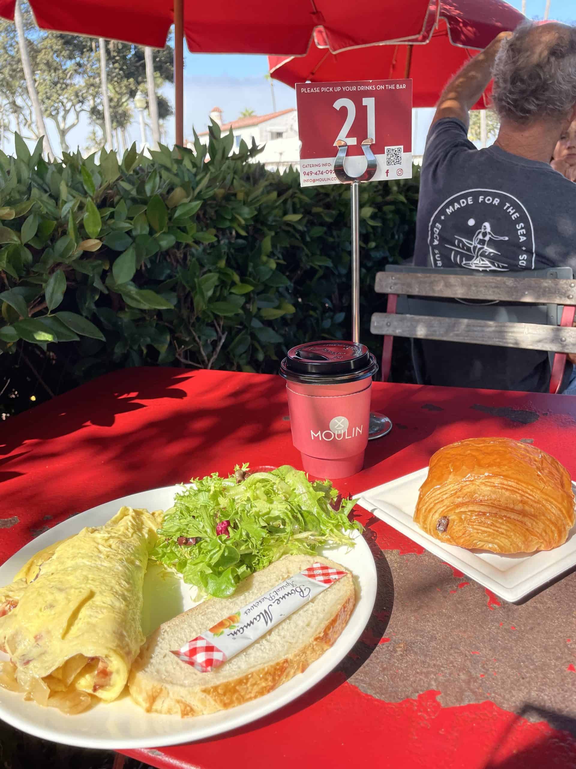 an omelette with salad and bread on a plate, a coffee in a red to go cup and a chocolate croissant on a red table outside