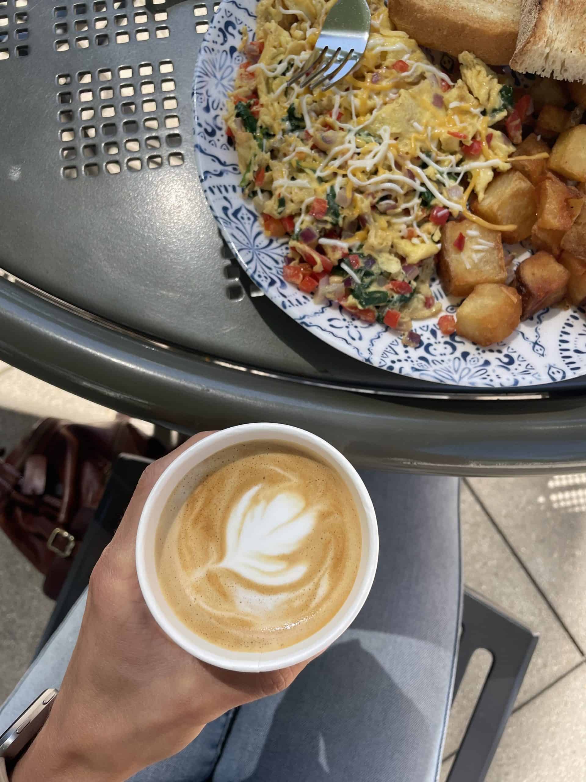 hand holding a paper cup with a latte in it near a metal table to a breakfast plate on it