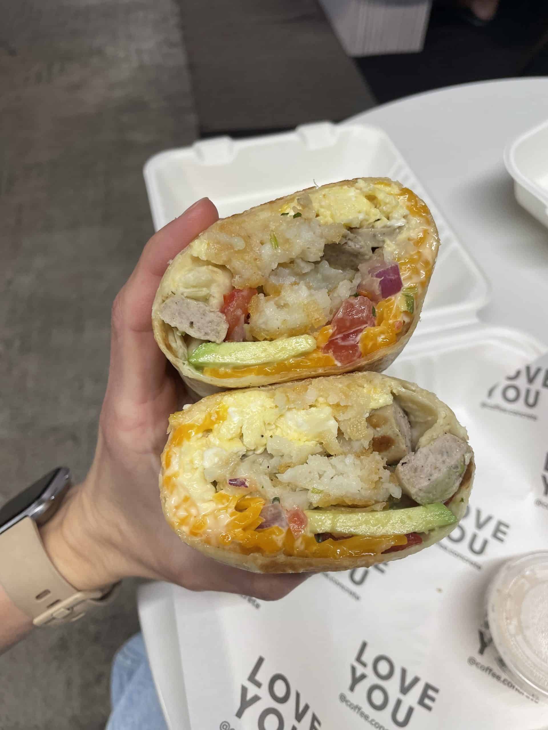 someone holding a breakfast burrito cut in half with egg, cheese, avocado, hash browns and tomatoes inside